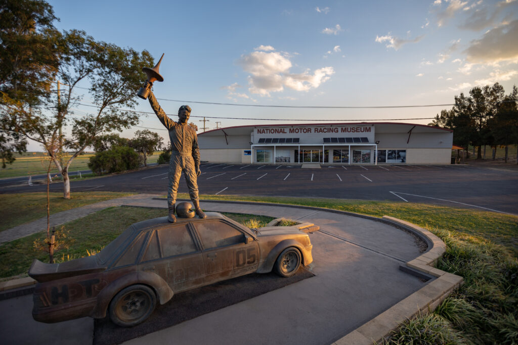 statue of a man standing on a racecar outside a museum building at golden hour