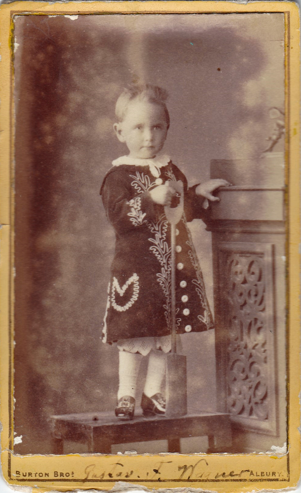 aged photograph of a small child standing on a stool and gripping carved wood furniture for support, he wears an embroidered coat buttoned up