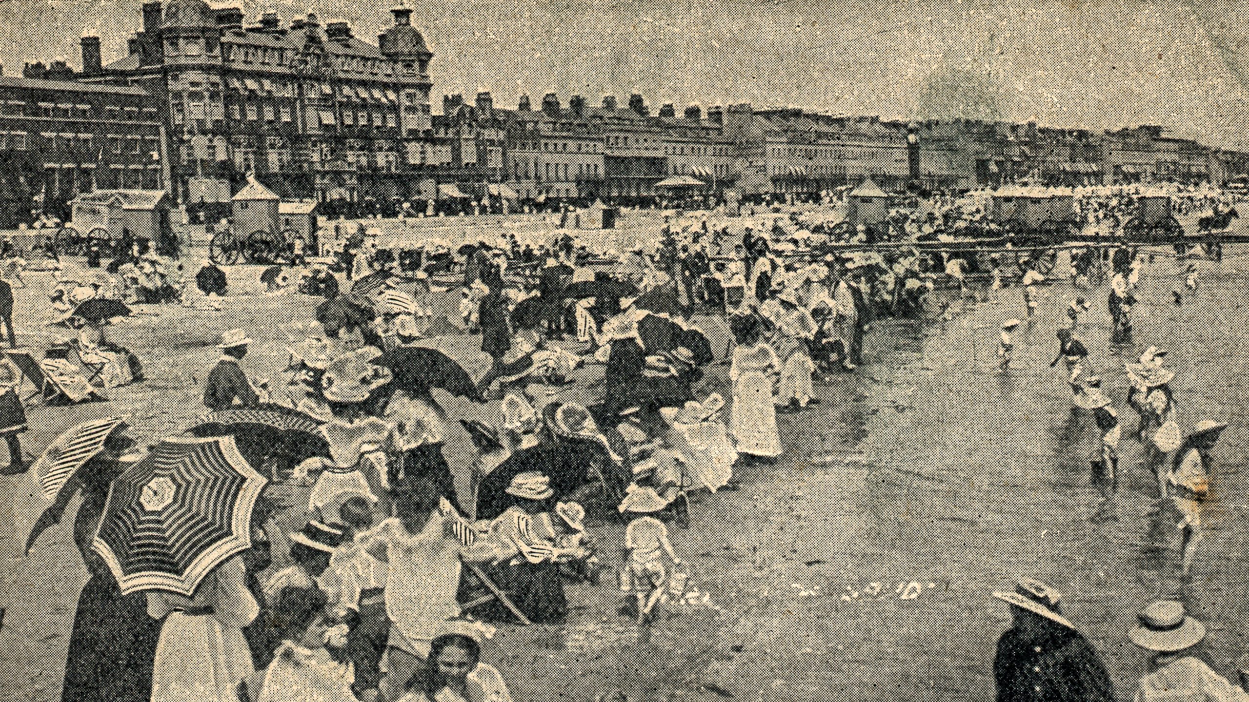 black and white postcard with image of packed beach with people in early twentieth century fashion, many hold parasols