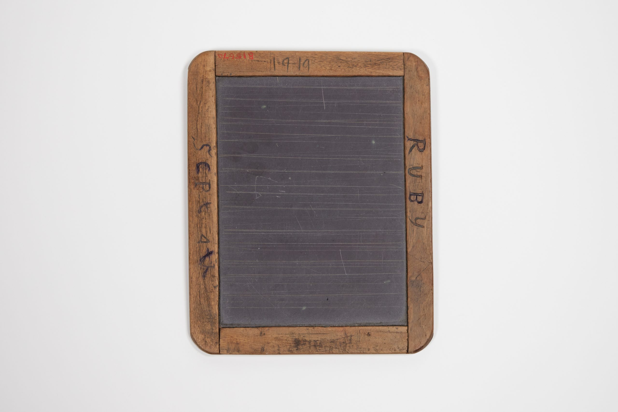 Black writing slate which has a wooden frame with curved corners. The frame is inscribed in handwriting saying "RUBY," "1919," and "SCP"