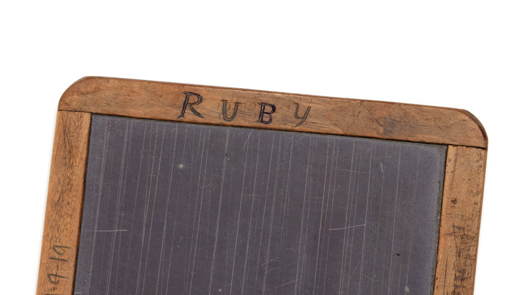 Close up of a black writing slate which has a wooden frame with curved corners. The frame is inscribed in handwriting saying "RUBY"