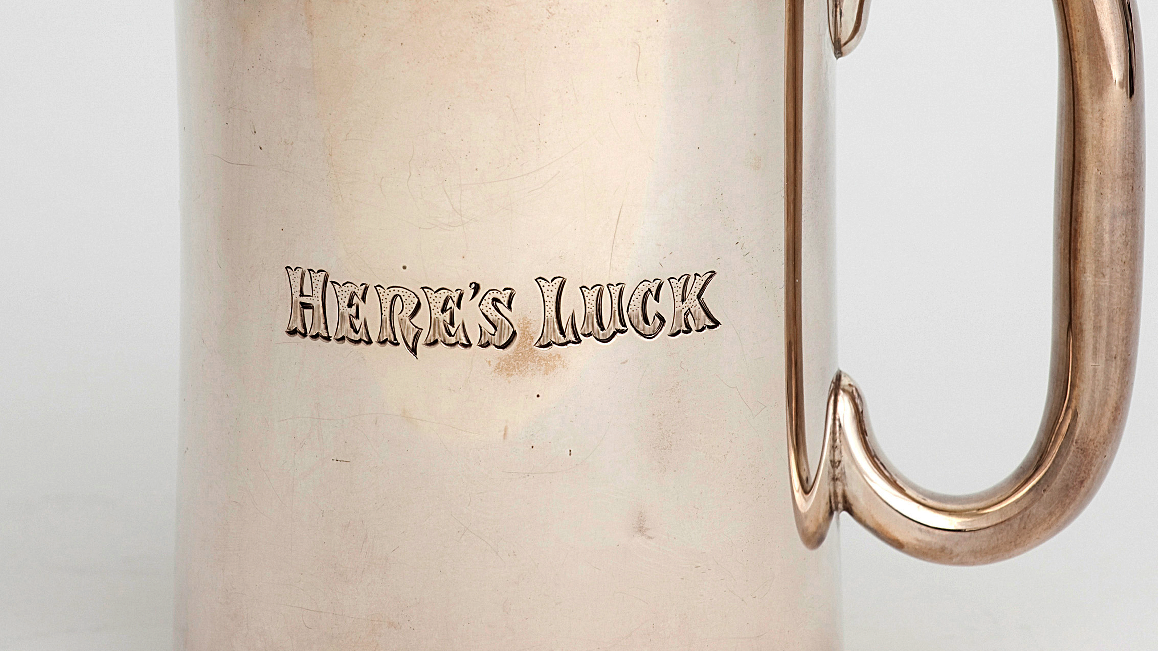 Close up of simple silver mug, slightly oxidised, with an inscription "HERE'S LUCK"