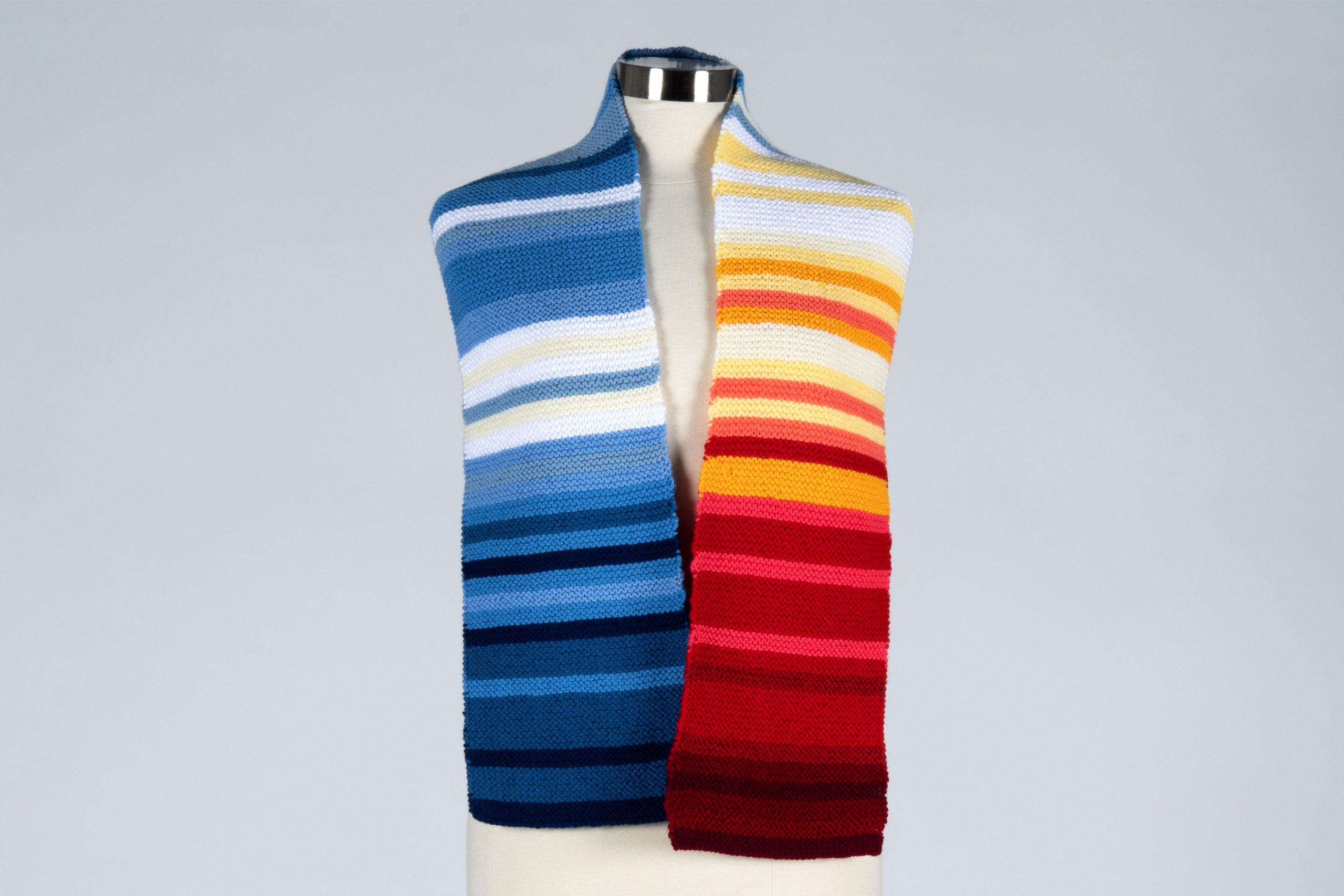 Mannequin wears a striped scarf around the shoulders. One side has various shades of blue and white while the other has various shades of red and white.