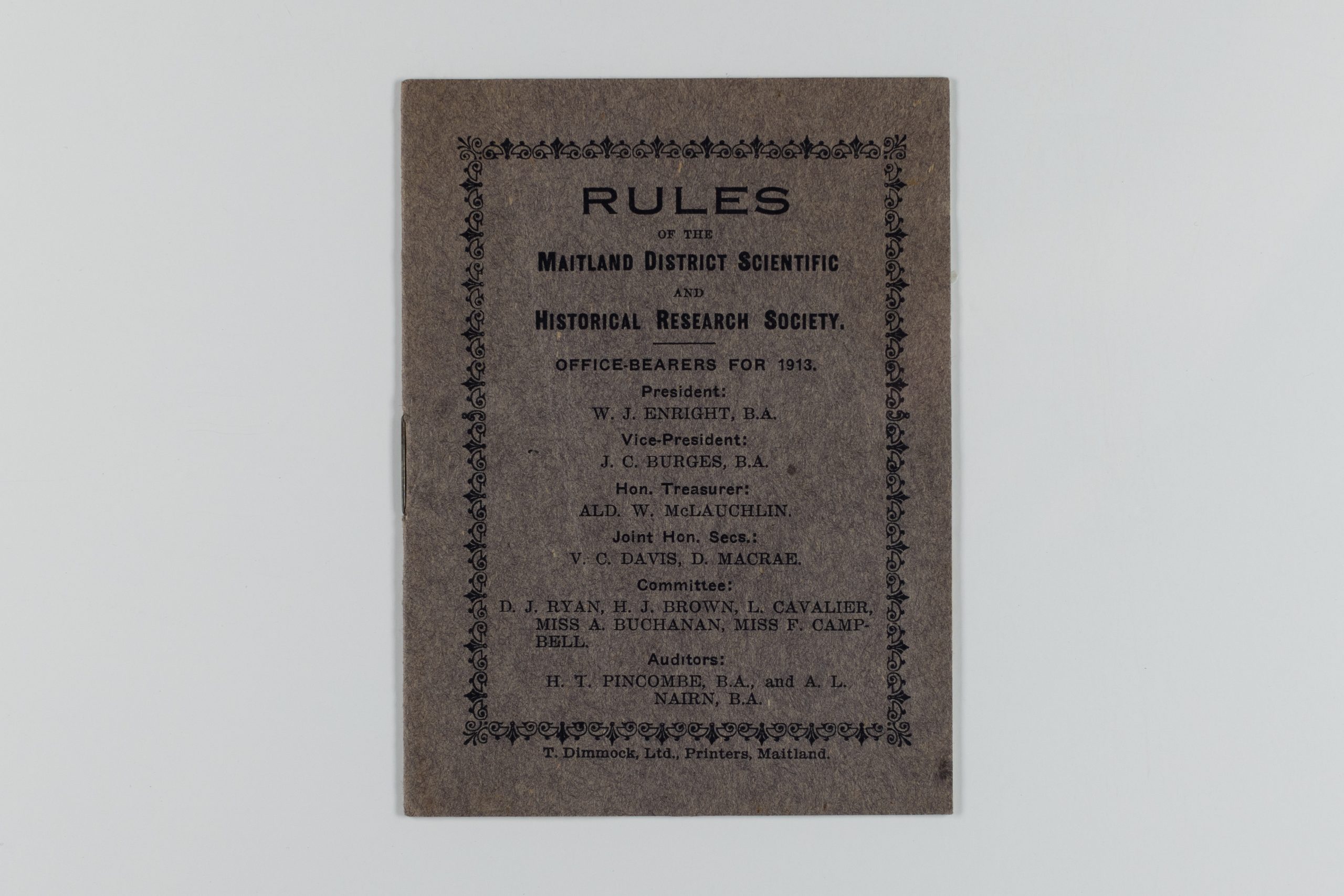 Front cover of brown, slightly tarnished book. It reads: "RULES OF THE MAITLAND DISTRICT SCIENTIFIC AND HISTORICAL RESEARCH SOCIETY. OFFICE-BEARERS FOR 1913. President: W.J. Enright, B.A. Vice-President: J.C. Burges, B.A. Hon. Treasurer: ALD. W. McLauchlin. Joint Hon. Secs: V.C. Davis, D. Macrae. Committee: D.J. Ryan, H.J. Brown, L. Cavalier, Miss A. Buchanan, Miss F. Campbell. AuditorsL H.T. Pincombe, B.A., snd Nairn, B.A. T. Dimmock, Ltd., Printers, Maitland"