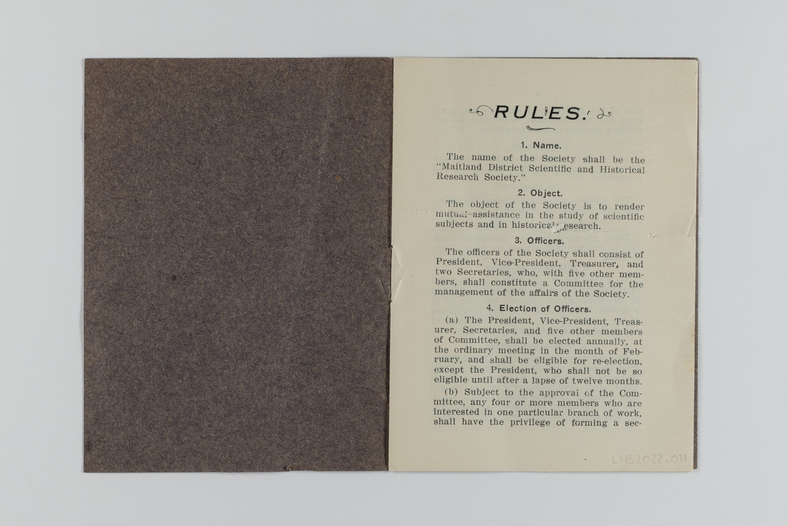Printed book with brown cover opened to the first page. It showcases rules 1-4, including the name and objective of the society, as well as the role of officers and their election.