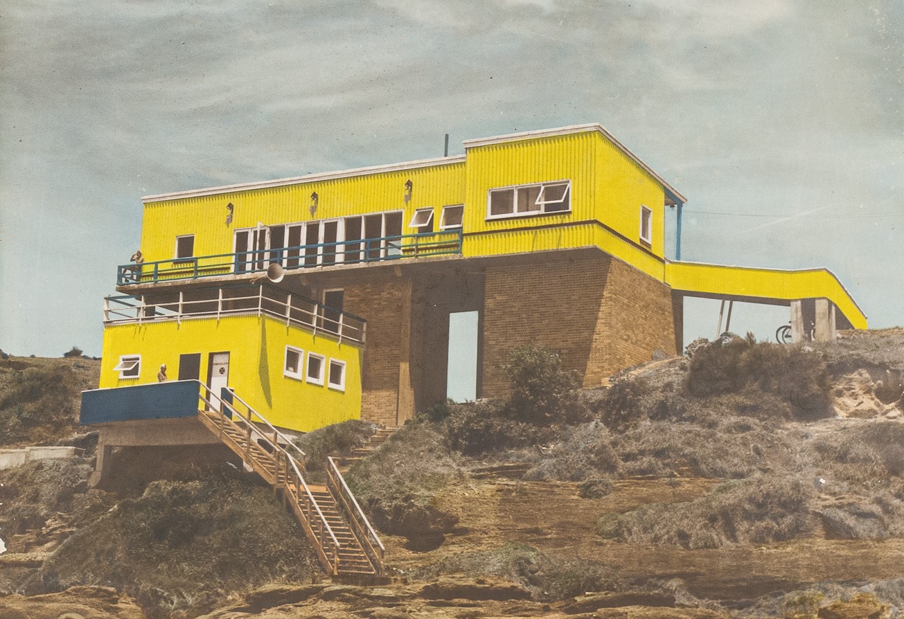 Black and white photograph, hand-tinted to appear with colour, of the Caves Beach club house in 1959. It is a yellow two-storey building on the bushy area sloping towards the beach, stairs extend from up from the sand into the lower floor.