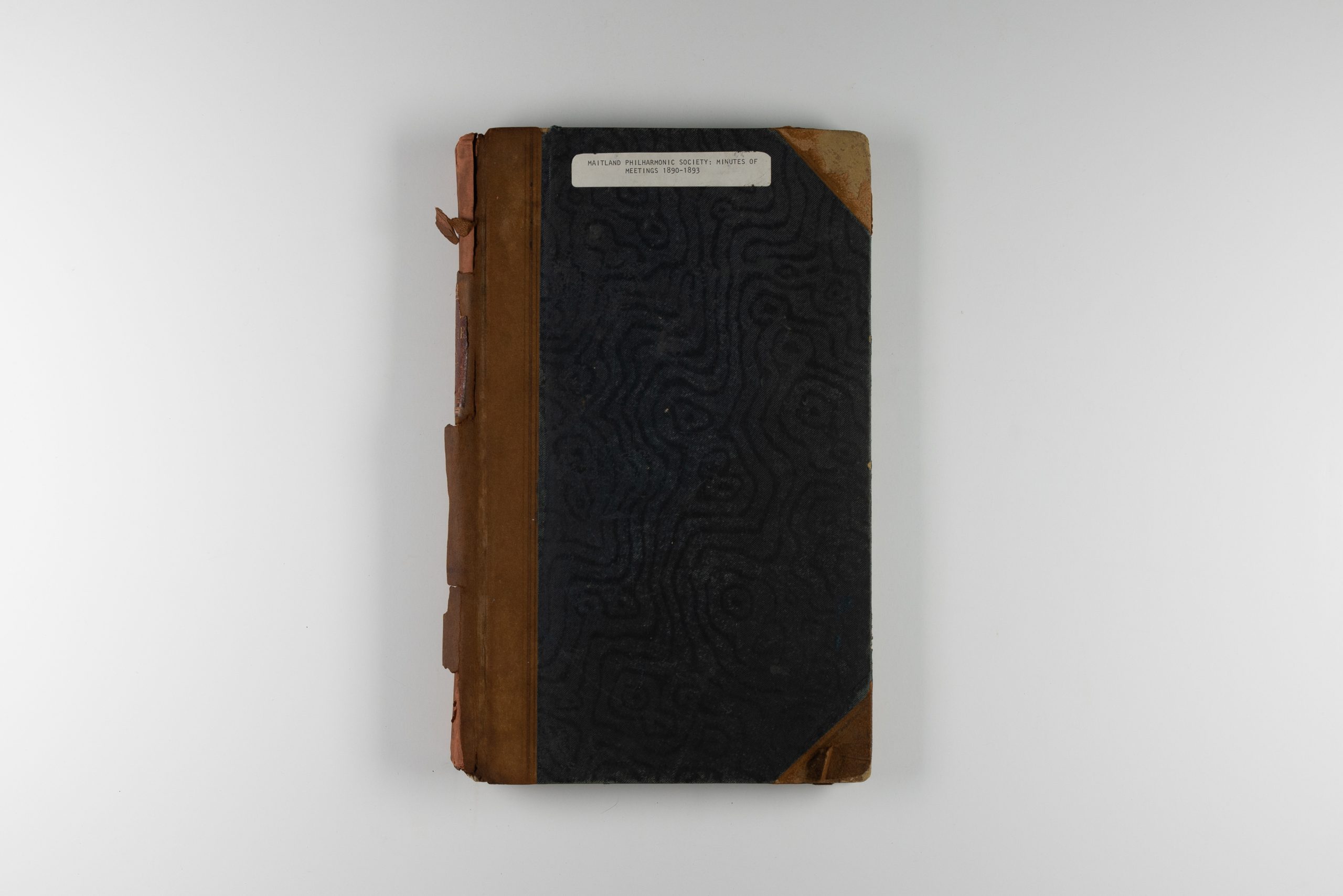 Front cover of a book, the cover is black with brown leather strips on the spine and corners. A thin, white, printed label is affixed to the top, it reads: "MAITLAND PHILHARMONIC SOCIETY: MINUTES OF MEETINGS 1890-1893"