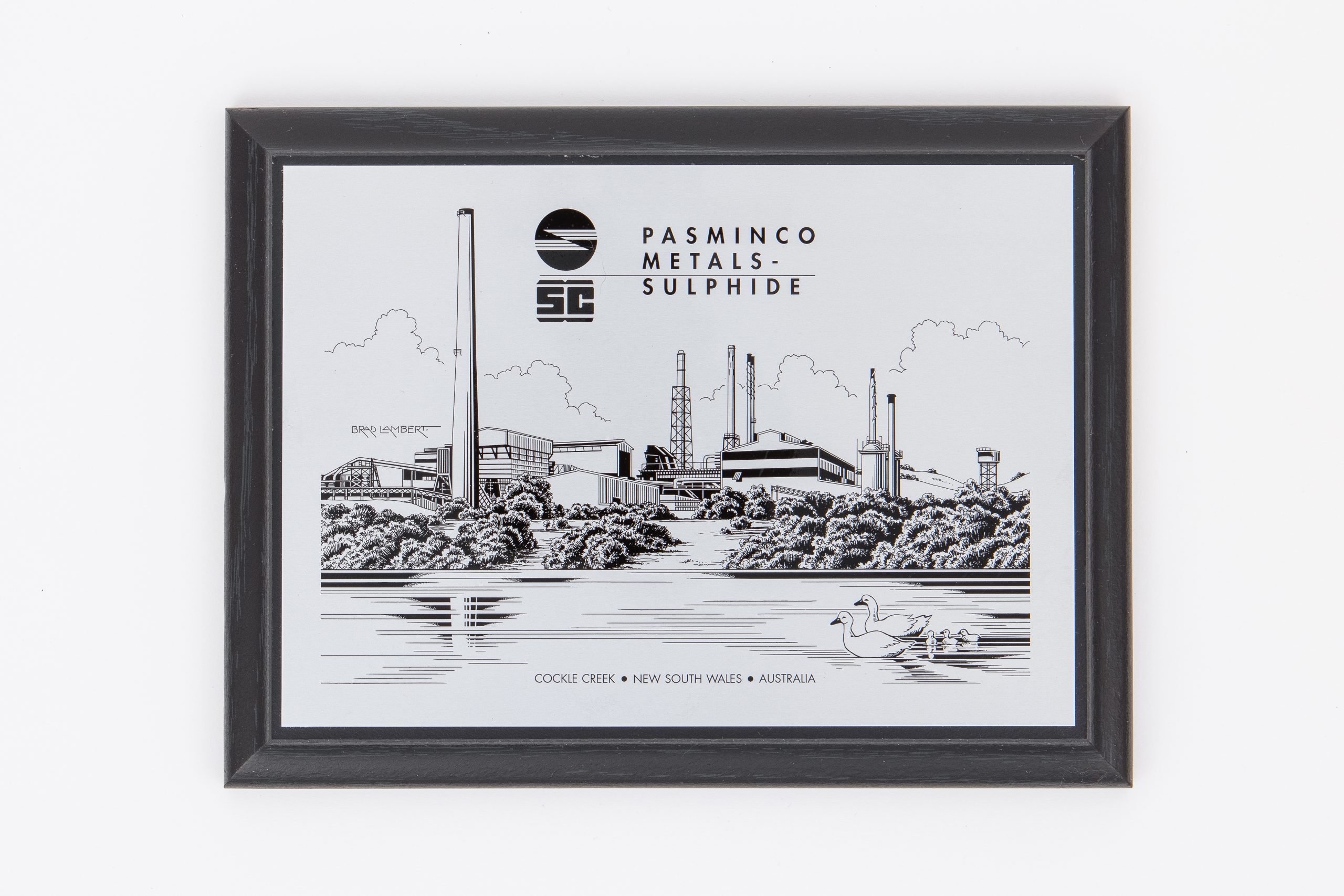 MDF plaque with black frame. It depicts a smelter beneath a relaxed, cloudy sky, behind a body of water where a family of ducks swim.