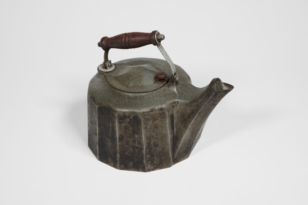 Black metal kettle with round but angular shape, akin to a hexagon but with more sides; wooden handle is attached to the top while the spout is simarly angular and pointed