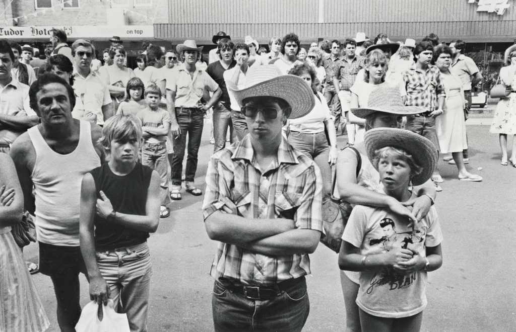 Black and white photograpg of a large group of people standing in the street facing towards the camera. People of all ages wear cowboy hats and plaid.