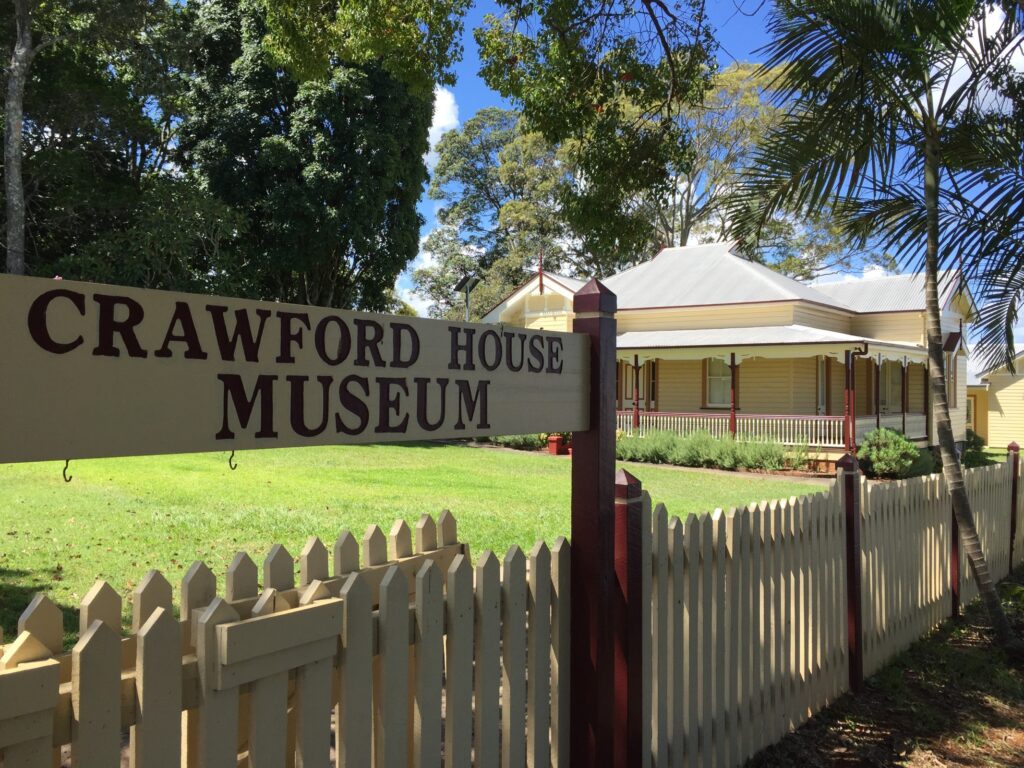 sign reading 'Crawford House Museum' in front of a single-storey historic house with beige walls and fenced verandah