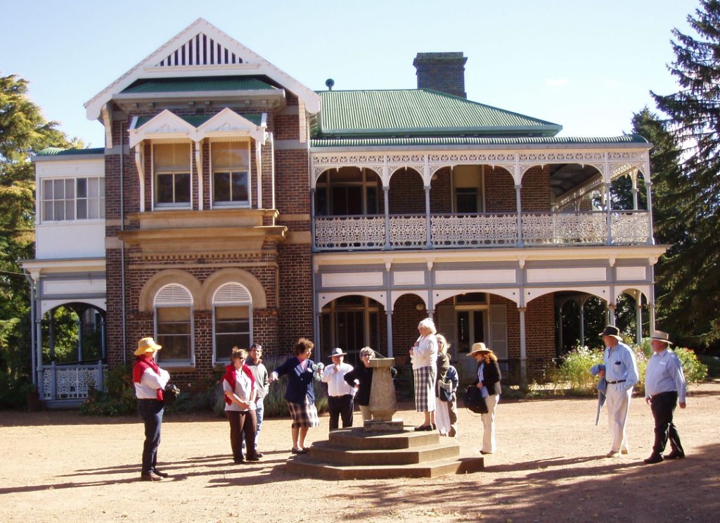 a group of people stand around a drinking fountain in front of a two-storey historic brick house with a green roof and cream-coloured adornments to the verandah
